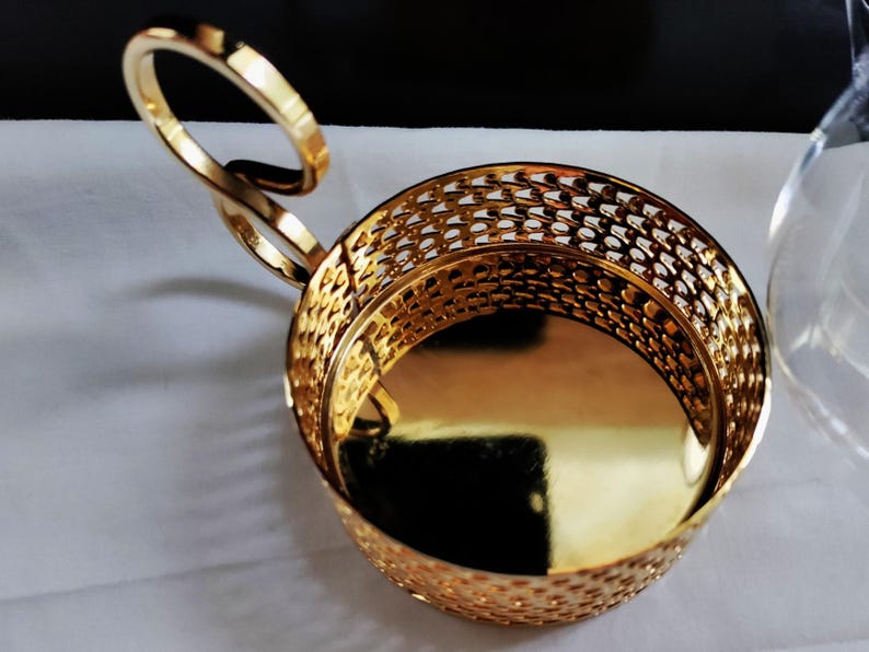 Puede incluir: Un portavasos de metal dorado con una base s&oacute;lida pulida y una pared lateral con un patr&oacute;n de celos&iacute;a decorativo. El soporte cuenta con un asa &uacute;nica en forma de bucle curvo que se extiende desde el lateral, dando una silueta distintiva.