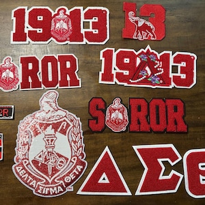 Peut inclure: Divers écussons brodés rouges et blancs, avec des lettres grecques, le chiffre 1913 et des écussons de sororité. Certains écussons incluent le mot "SOROR" et un éléphant. Ces écussons sont probablement pour vêtements ou accessoires.