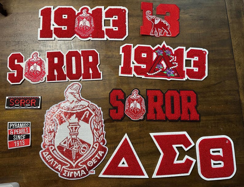 May include: A collection of red and white embroidered patches, featuring Greek letters, the number 1913, and sorority crests. Some patches include the word "SOROR" and an elephant graphic. The patches are likely for apparel or accessories.