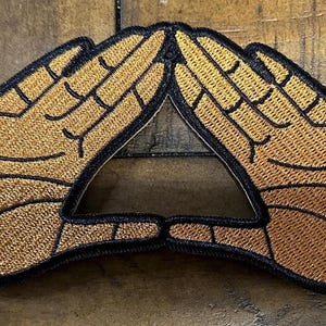 May include: Embroidered patch featuring two hands forming a triangle. The hands are tan with black outlines, and the patch has a black border. The design is a symbol of unity and connection.