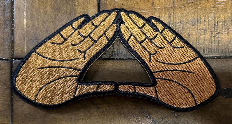 May include: Embroidered patch featuring two hands forming a triangle. The hands are tan with black outlines, and the patch has a black border. The design is a symbol of unity and connection.