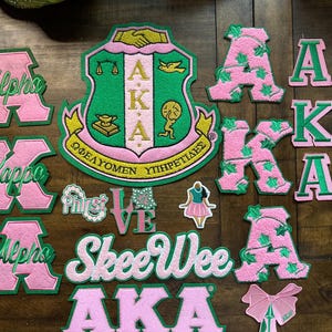 May include: A collection of pink and green embroidered patches and appliques, including letters spelling "Alpha Kappa Alpha", a crest, and other decorative elements. The patches are arranged on a wooden surface.