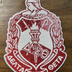 May include: Red and white sequined patch featuring the Greek letters Delta, Sigma, and Theta, with a torch and a figure in a helmet. The patch has a shield shape with a white border and the text "Delta Sigma Theta".