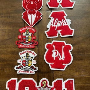 May include: A selection of red and white embroidered patches. Designs include a rabbit in a tuxedo, Greek letters, and crests with text such as "Kappa Alpha Psi" and "Fraternity, Inc." The number "1911" is also featured.