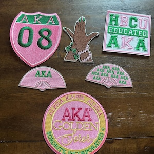 May include: A collection of embroidered patches in pink, green, and gold. The patches feature the Greek letters AKA, a hand with a pearl necklace, and the words "HBCU EDUCATED AKA". Other patches include a shield with "AKA 08" and fan shapes.