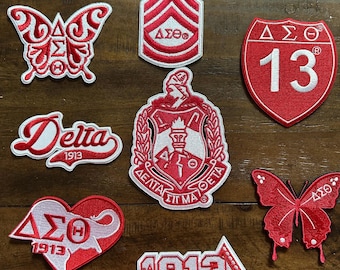 May include: A selection of red and white embroidered patches in various shapes, including butterflies, shields, and badges. Some feature the Greek letters Delta Sigma Theta and the number 13. The year 1913 is also displayed on some patches.