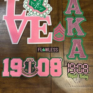 Può includere: Una collezione di toppe ricamate rosa e verdi. Le toppe includono la parola "LOVE", le lettere "AKA", i numeri "1908" e la parola "FLAWLESS". Alcune toppe hanno accenti glitterati.