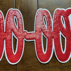 May include: A red sequined patch with the text "GO-OUP" in a cursive font. The letters are outlined in white. The patch is on a brown wooden surface.