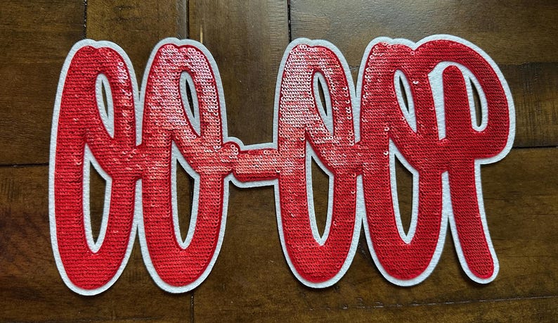 May include: A red sequined patch with the text "GO-OUP" in a cursive font. The letters are outlined in white. The patch is on a brown wooden surface.