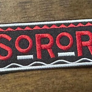May include: A rectangular patch with the word "SOROR" embroidered in red on a black background. The patch has a white border and red decorative elements. The patch is likely for clothing or accessories.