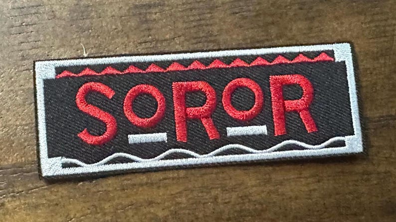 May include: A rectangular patch with the word "SOROR" embroidered in red on a black background. The patch has a white border and red decorative elements. The patch is likely for clothing or accessories.