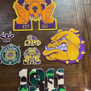 May include: Embroidered patches with a bulldog mascot in orange, purple, and yellow. Some patches include the text "EST. 1911" and "Omega Delta Phi". The patches are in various shapes and sizes, suitable for clothing or accessories.