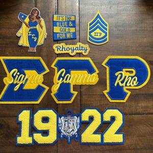 May include: A selection of blue and gold embroidered patches, featuring Greek letters, a figure, and text. The patches display the Greek letters Sigma, Gamma, and Rho, alongside the year 1922 and the phrase "It's the Blue & Gold for Me."
