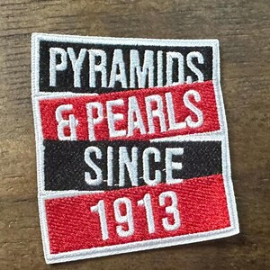 May include: Embroidered patch with the words "PYRAMIDS & PEARLS SINCE 1913" in white lettering. The patch has a rectangular shape with a black, red, and white color scheme. The text is stacked in three rows.