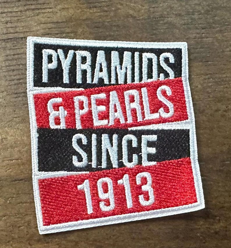May include: Embroidered patch with the words "PYRAMIDS & PEARLS SINCE 1913" in white lettering. The patch has a rectangular shape with a black, red, and white color scheme. The text is stacked in three rows.