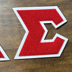 May include: Three red and white embroidered patches in the shape of a triangle, a Greek letter Sigma, and a letter O. The patches have a textured, felt-like appearance and are set against a wooden background.