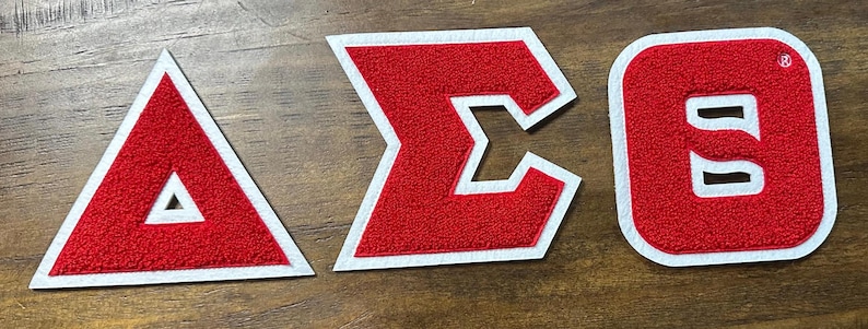 May include: Three red and white embroidered patches in the shape of a triangle, a Greek letter Sigma, and a letter O. The patches have a textured, felt-like appearance and are set against a wooden background.