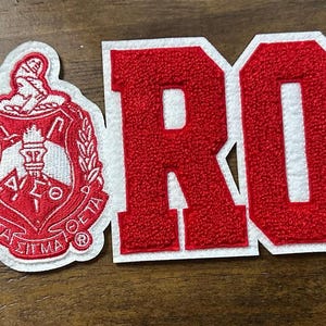 May include: Red and white embroidered patches spelling out "SOROR" with a Greek sorority crest. The letters are red with a white border. The crest is red and white with Greek letters.