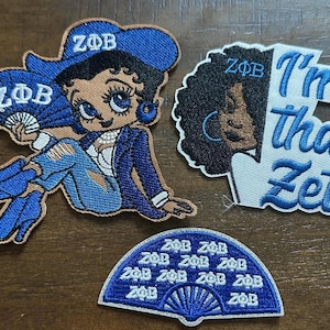 Könnte beinhalten: Gestickte Aufnäher mit Designs der Zeta Phi Beta-Verbindung. Ein Aufnäher zeigt eine Cartoon-Frau in blauer Kleidung, ein anderer lautet "I'm that Zeta", und ein dritter zeigt einen Fächer mit den Buchstaben der Verbindung.