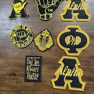 May include: A collection of black and gold embroidered patches featuring various designs including the letter A, a hand gesture, a crown, a pharaoh mask, and the text 'Who Ya Wit', 'Alpha Phi Alpha', 'Alpha', and 'Did The Right Thing' in a rectangular shape.