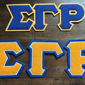 Pode incluir: Dois conjuntos de patches de letras gregas, que soletram "ΣΓΡ". Um é azul com borda dourada, o outro é dourado com borda azul. Os patches são feitos de um material texturizado, provavelmente feltro ou chenille, em uma superfície de madeira.