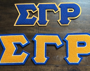 May include: Two sets of Greek letter patches, each spelling "ΣΓΡ". One set is blue with gold trim, the other is gold with blue trim. The patches are made of a textured material, likely felt or chenille, and are displayed on a wooden surface.