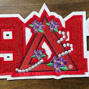 May include: A red and white embroidered patch with the numbers "1913". The patch features a red triangle with purple flowers and a string of pearls. The patch is on a wooden surface.