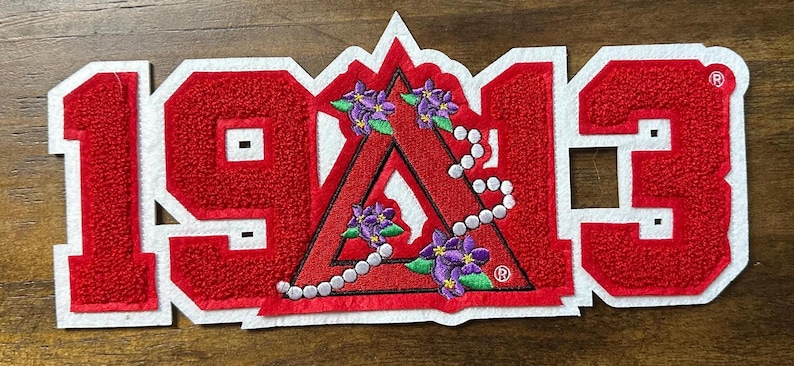 May include: A red and white embroidered patch with the numbers "1913". The patch features a red triangle with purple flowers and a string of pearls. The patch is on a wooden surface.