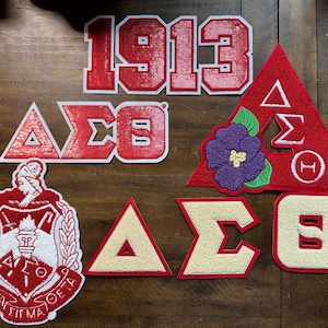 May include: A collection of red and white embroidered patches, including the number "1913", Greek letters, and a crest. Some patches feature a triangle shape, while others have a square or shield design. A purple flower is also featured.
