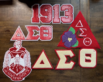 May include: A selection of red and white embroidered patches, featuring the number "1913", Greek letters, and a crest. Some patches are triangular, while others are square or shield-shaped. A purple flower is also present.
