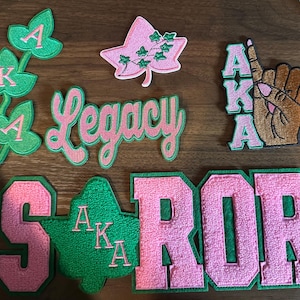 Puede incluir: Colección de parches bordados en rosa y verde. Los parches incluyen las letras AKA, una mano con el dedo índice levantado, hojas de hiedra y la palabra "Legacy".