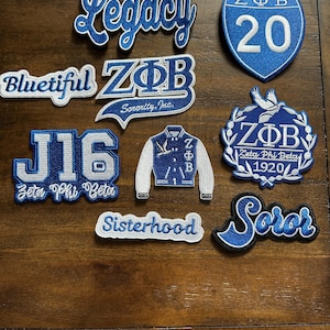 May include: A collection of blue and white embroidered patches with various designs and text. The patches include the words "Legacy", "Bluetiful", "Sisterhood", and "Soror". Other patches feature "ZΦB", "J16", and a jacket design.
