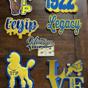 Könnte beinhalten: Eine Sammlung von gestickten Aufnähern in Gelb, Blau und Weiß. Designs umfassen die Zahl "1922", das Wort "Legacy", einen Pudel und das Wort "Love" mit einem Pudel und der Zahl "1922". Weitere Aufnäher zeigen griechische Buchstaben und Phrasen.