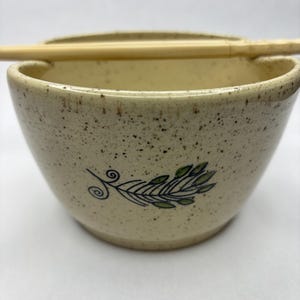 May include: A speckled ceramic bowl with a green leaf design and a bamboo chopstick resting on the rim. The bowl has a cream-coloured interior and a speckled exterior. The leaf design is hand-painted in green and black.