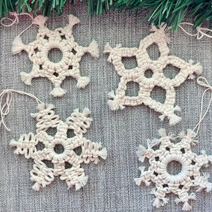 May include: Four handmade, off-white crochet snowflake ornaments with fringe details. Each snowflake has a loop for hanging. The ornaments are displayed on a gray surface, with evergreen branches in the background.