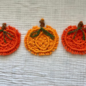 May include: Three crocheted pumpkin coasters in orange and yellow with green stems. The coasters are on a white fabric background.