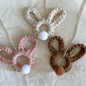 May include: Three crocheted bunny rabbit shapes with white pom-poms. The bunnies are in white, pink, and brown.