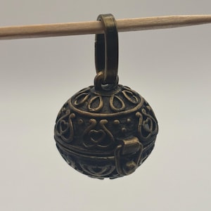 May include: A spherical bronze-toned metal pendant featuring an intricate embossed pattern of hearts and circular motifs. The charm includes a small hinged latch mechanism on the side and a sturdy loop at the top for attachment to a chain or cord.
