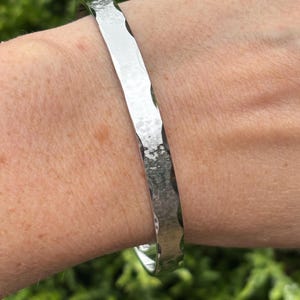 May include: A silver-colored metal bangle bracelet featuring a hammered texture on the outer surface. The bracelet has a flat, polished profile with distinct indentations along the edges, creating a reflective and textured appearance on the wrist.