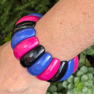 May include: A stretch bracelet composed of smooth, glossy, rounded segments in alternating colors of hot pink, black, and royal blue. The segments are arranged in a repeating pattern, creating a continuous, flexible band around the wrist.