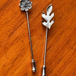 May include: Two silver-toned lapel pins. One pin features a detailed flower design, and the other has a leaf shape. Both pins have long, slender stems and decorative bases. The pins are displayed on a warm, wooden surface.