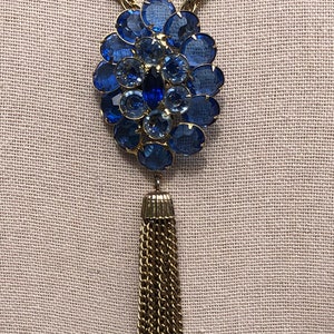 May include: A gold chain necklace with a large blue rhinestone pendant and a gold tassel.