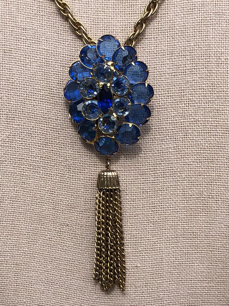 May include: A gold chain necklace with a large blue rhinestone pendant and a gold tassel.