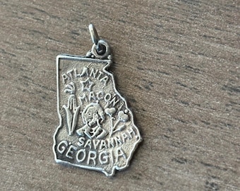 Könnte beinhalten: Silberfarbener Anhänger in Form des Bundesstaates Georgia. Der Anhänger trägt die Namen Atlanta, Macon und Savannah. Er zeigt dekorative Elemente wie einen Stern, Blumen und eine stilisierte Figur.