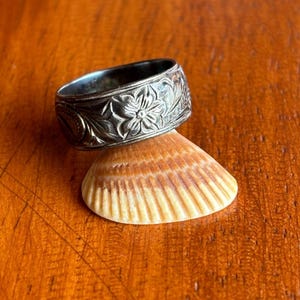 May include: A silver ring with a floral pattern rests on a seashell. The ring has a dark, oxidized finish, and the floral design is intricately carved around the band. The seashell is light brown with a textured surface.