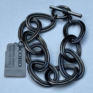 May include: A black metal bracelet with a toggle clasp. The bracelet is made of linked oval shapes with a textured surface. The tag attached to the bracelet reads "CORO French Gray Collection".