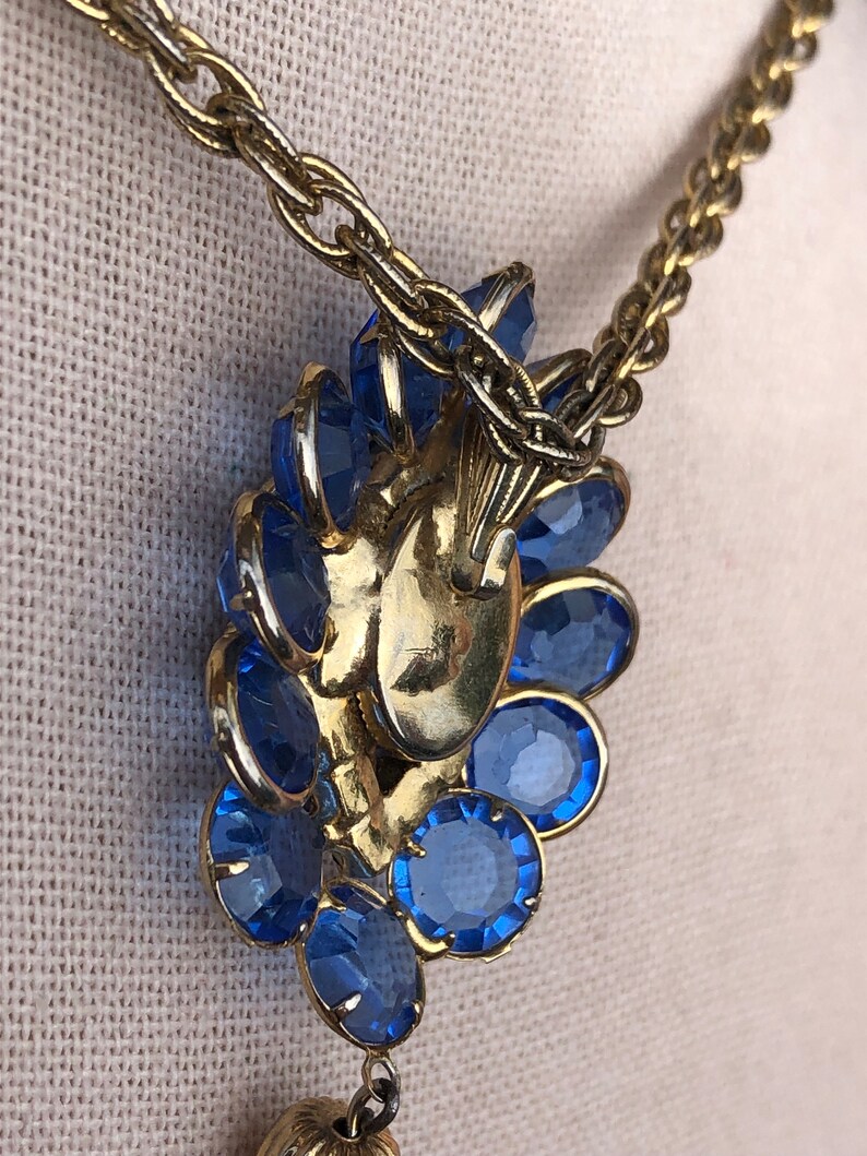 May include: A gold chain necklace with a large blue rhinestone pendant. The pendant is shaped like a flower and has a dangling drop.