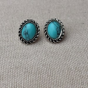 May include: A pair of oval turquoise stud earrings. Each earring features a vibrant blue-green stone set within a silver-toned, rope-like border. The earrings are displayed on a textured, neutral-colored surface.