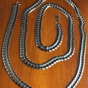 May include: A silver-colored necklace with a unique, textured design. The necklace is made of interlocking metal pieces, creating a flexible and eye-catching chain. The necklace is displayed on a wooden surface.