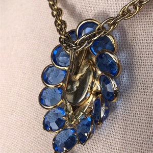 May include: A gold chain necklace with a blue rhinestone pendant and a gold tassel. The pendant is shaped like a flower with multiple layers of rhinestones.